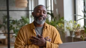 A middle-aged man with a short gray beard sits indoors with his eyes closed and both hands crossed over his chest, conveying calm and gratitude. He wears a mustard-colored shirt over a gray tee. Soft natural light, large windows, and green houseplants in the background create a peaceful, reflective atmosphere, with a laptop visible in front of him.