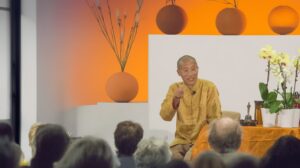 Master Mingtong, wearing a gold traditional-style robe sits at the front of a room, speaking and gesturing toward an audience seated in front of him. The setting is calm and ceremonial, with an orange-and-white backdrop, minimalist round vases, branches, and a table holding orchids and small ritual objects.