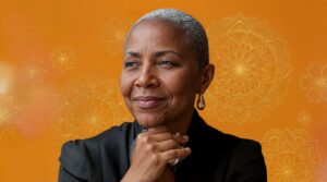 An older woman with closely cropped gray hair rests her chin on her hand and looks off to the side with a thoughtful, gentle smile. She wears a dark jacket and small hoop earrings. The background is a warm orange tone decorated with delicate, mandala-like floral line patterns, creating a calm, reflective atmosphere.