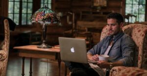 A man sits in a cushioned armchair inside a rustic, wood-paneled room, working on a laptop balanced on his lap while holding a mug. A small wooden side table beside him holds a decorative stained-glass lamp and a book, with soft daylight filtering through windows in the background, creating a quiet, cozy atmosphere.